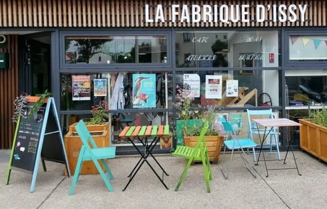 Photo de la facade de La Fabrique d'Issy. La façade est composée d'une grande baie vitrée sur laquelle on peut voir plusieurs affiches. Des chaises et des tables de récupération sont posées devant la façade à côté de bacs de fleurs.