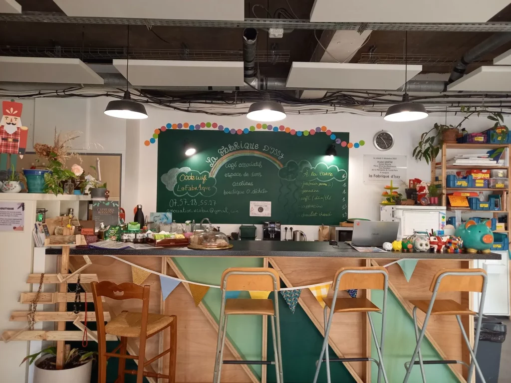 Photo du bar de La Fabrique d'Issy devant lequel sont posées trois grande chaise du même modèle ainsi qu'une plus petite chaise. Le tout provenient de dons. Derrière le bar est situé un grand tableau avec les coordonnées de la Fabrique, le nom du lieu dessiné au dessus d'un arc-en-ciel ainsi que la liste des boissons.